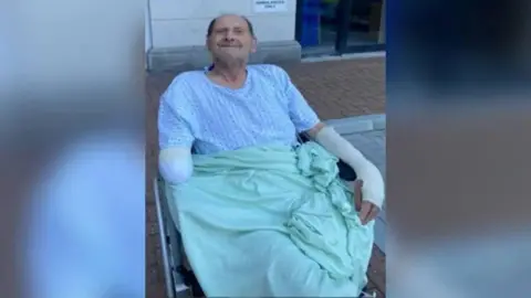 Sussex Police Vic Franklin smiling in a wheel chair while wearing a light blue blanket and a hospital gown. He is a elderly white man who is bald on top with dark hair on the sides of his head. He has a bandage on his right arm which has been amputated by the elbow and a bandage on his left arm and hand.