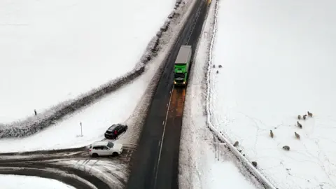 An aerial picture of a road with a truck and several cars driving on it. An intersecting roads has cars waiting to turn onto the road on it. The whole area, apart from the cleared roads, is covered in snow. Sheep are seen in a nearby snow-covered field. 