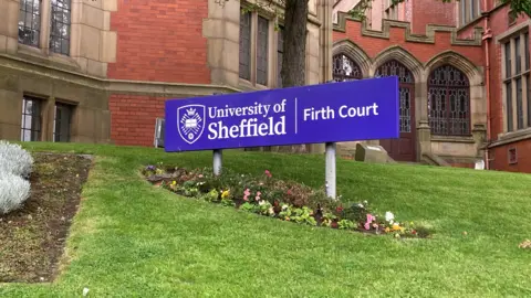 A purple sign says The University of Sheffield, Firth Court. It is on a grass verge with a red-brick building in the background.
