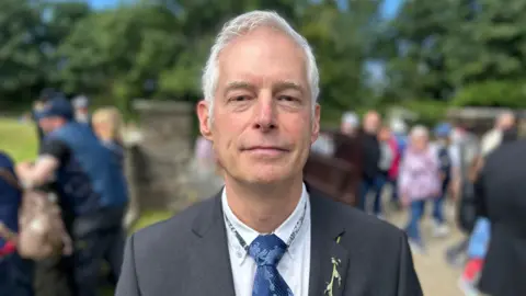 Chris Pay wears a navy blue suit and tie with a white shirt. He has short grey hair 