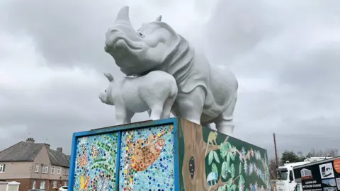 A fibreglass statue of a rhino with a baby on top of a decorated plinth in front of a housing estate on a grey day