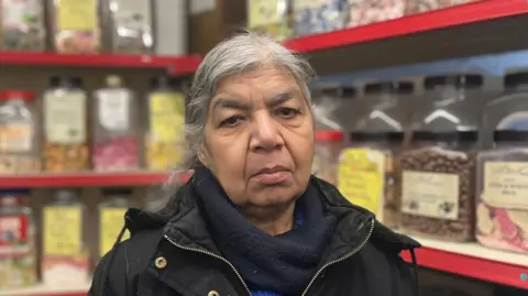 BBC Jagdish Kaur, stood in front of jars of sweets, looking at the camera