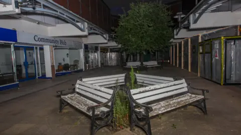 Shrewsbury From Where You Are Not A dark passageway at the shopping centre between shops with a quarter of benches arranged around a flowerbed, growing weeds. The benches are in a diamond shape together and have white, peeling paint with black stands at either end.