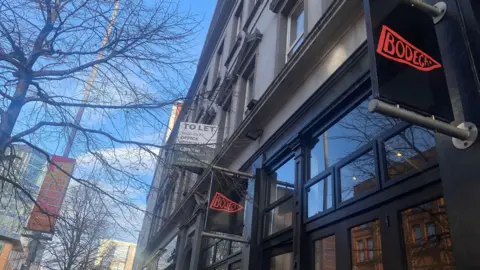 Exterior of Bodega Bagels. The shop is painted black with black signs and a red logo. The building above is grey. There are trees and a blue sky.