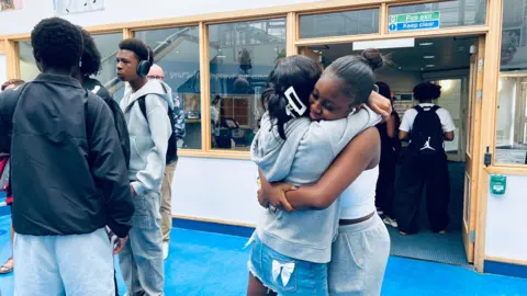 Two girls are hugging each other in a school hall, next to a group of boys.