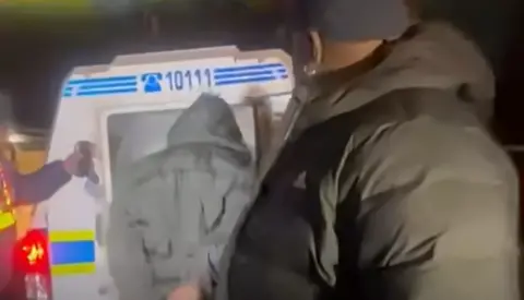 SABC NEWS A man in a puffy coat and dark-coloured baseball cap is in the foreground, but looking away from the camera, towards someone in a grey coat being led into a police van. It is dark.