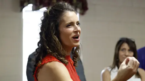 Julia Letlow is seen talking to constituents which smiling and appearing to talk. She is turned to the side and is seen from the shoulder up. She is wearing a red top.