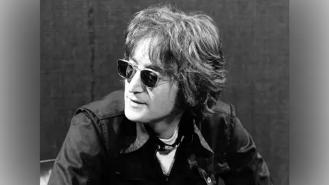 John Lennon, with medium-length brown hair, wearing dark sunglasses and a jacket sits and speaks to someone off camera 