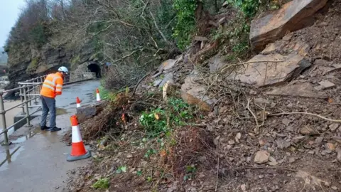 A man wearing an orange high vis jacket standing next to a landslide with broken rocks and trees 