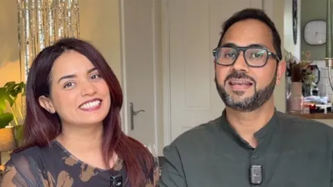 A woman and a man with their arms linked, seated and smiling at the camera