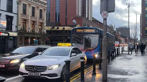 A white taxi with a yellow Value Cabs sign on the roof drives in a bus lane on Great Victoria Street. There is a blue bus behind it, also in the bus lane. Text on the bus display says Merry Christmas. There is a black car in the lane beside the taxi. Pedestrians walk down the street.