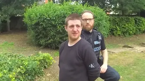 Thames Valley Police Robert Richens is kneeling down in handcuffs with a police officer behind him.