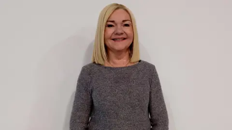 Paula Gibson is standing against a white wall. She's wearing a grey jumper and has a chain under her jumper.