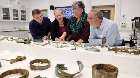 Durham University Two men and two women gather around a long table to look at the artefacts spread across it