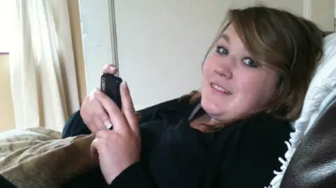 Family handout Sophie Alderman, who has shoulder length brown hair and is smiling at the camera. She is lying on a sofa indoors while holding phone in her hands and wearing a black, long-sleeved top.