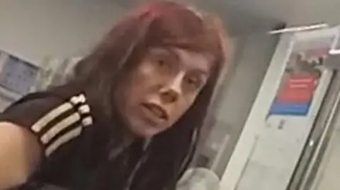 Devon and Cornwall Police Holly Collinson captured on CCTV. She is looking into the camera and has dark red hair. She is wearing a black t-shirt with white stripes on the sleeves.