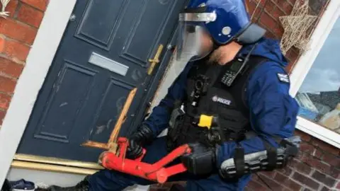 Greater Manchester Police A police officer dressed in navy blue wearing a police vest and a hard hat with a visor attachment. He stands by the front door of a house with a large red device which he is using to cut into the wooden door of the property. His face has been blurred.