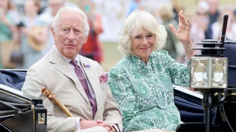 PA Media King Charles and Queen Camilla sitting in a carriage. The King is wearing a beige suit and holds a wooden walking stick. The Queen wears a green and white paisley print dress and is waving to the crowds.