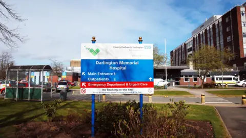 A blue and white sign outside Darlington Memorial Hospital which is a large, brick building to the right of the photo. The sign points to the main entrance and emergency department in red. Behind the sign is a patch of grass and car park.