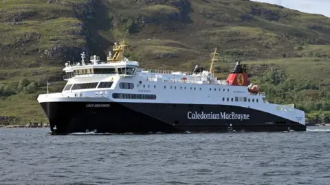 Loch Seaforth ferry. It is on the water with a hill behind it. The ferry is mainly white at the top with blue underneath. It has a red and black funnel and its name and Caledonian MacBrayne written in white on the side