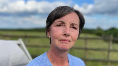 Older woman with dark hair wearing a light blue top standing in front of a fence