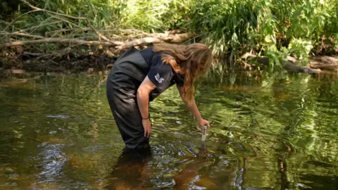 A woman in a black T-shirt and waders is checking the river temperature by putting a thermometer in the water 