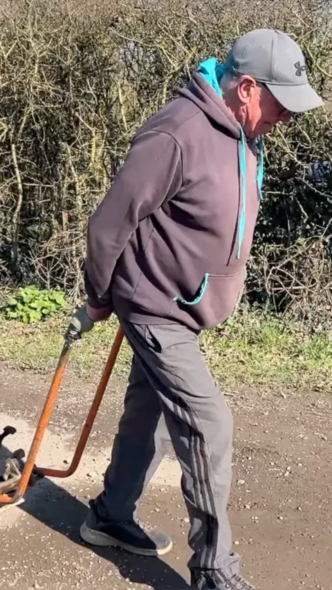 The image shows a person outdoors on a rural road, pulling a heavy whacker plate behind them. The individual is wearing a dark hoodie, dark trousers, gloves, and a cap. The whacker plate is being dragged along a rough, potholed surface.