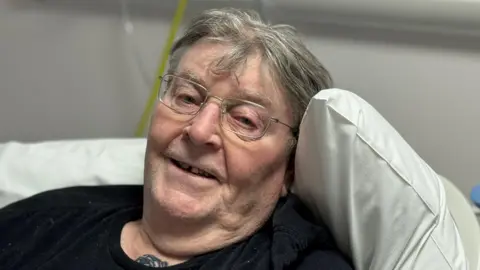 Ron Price is lying on a hospital bed and smiling. He has grey hair and glasses. He is wearing a dark top. 
