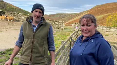 Daniel Wood is standing next to Sam Wood. Sam is a woman in her forties wearing a blue top with dark hair tied back. Daniel is wearing a jumper and a green jacket and blue woolly hat. Behind them is a stone wall with sheep up against it and in the distance are rolling hills on a sunny day.