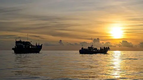 Two boats carrying lots of people float in a sea against a setting sun