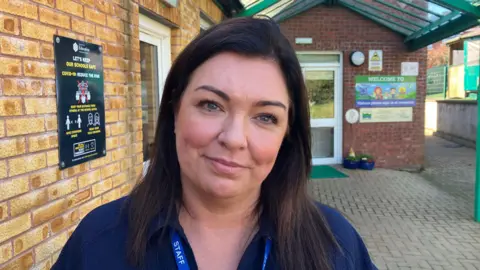 Evans has long dark brown hair and blue eyes. She is wearing a dark blue shirt and is standing outside in front of the entrance to the nursery.