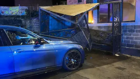 A blue car outside a pub with a smashed smoking shelter in front of the vehicle pinned against the wall of the building