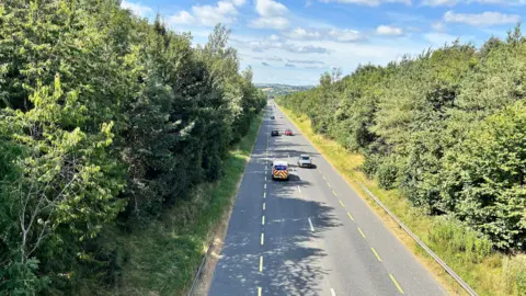 Vista aérea de un tramo de la carretera N2 cerca de Carrickmacross en un día soleado. Varios vehículos circulan por esta carretera nacional de un solo carril, bordeada de arcenes cubiertos de hierba y densa vegetación.