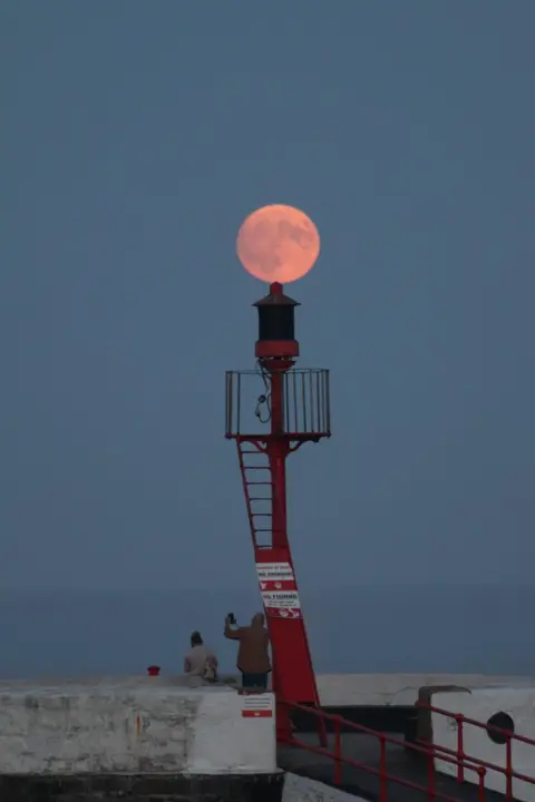 Andy Preston A red harbour navigation beacon stands on a pier in Looe, Cornwall, with a pink-orange full moon appearing directly above it and two people silhouetted nearby.