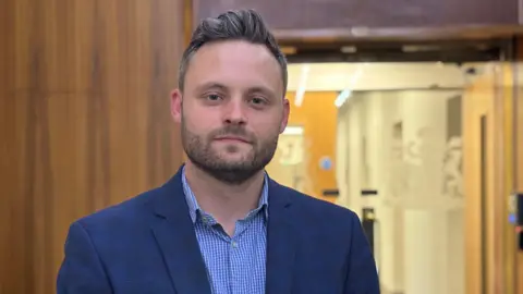 Former Nottinghamshire County Council leader Ben Bradley looks at the camera. He is wearing a blue and white open-collar shirt and dark blue jacket.