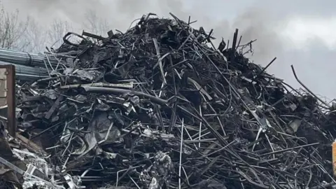WMFS A large pile of scrap metal which looks burnt. It is black and grey and there is smoke above it. 