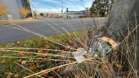 A bunch of flowers lie next to a tree, nearly buried under the dying stalks of grass. The tree is next to a road and beyond that are industrial buildings.