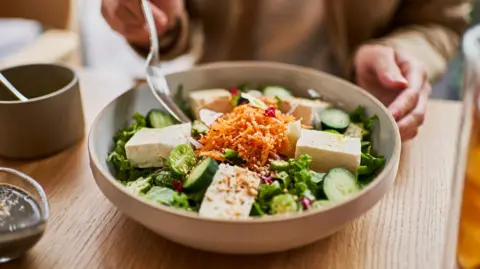 A stock image of a bowl of salad on a wooden table top. The salad contains grated carrot, tofu, cucumber and salad leaves. A person's hands, holding a fork, can be seen.