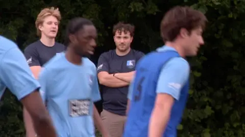Man watches footballer on sideline.