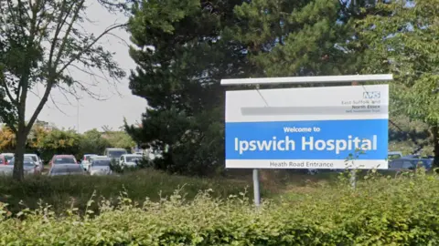 Google Maps A blue-and-white sign saying "Welcome to Ipswich Hospital". There is a hedge in front of it and trees behind it. A car park can also be seen in the background.