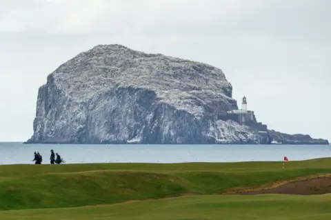 Chris Bell Large rocky island covered in seabirds rising from the sea, with a lighthouse perched on one side. In the foreground, a grassy golf course with a flag and several people walking along the links.