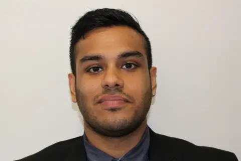 The Khalsa Academy A young man with short dark hair looks at the camera with a slight smile. he stands in front of a white background and wears a black suit jacket with a dark grey shirt.