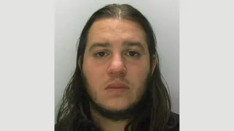 Gloucestershire Constabulary A police mugshot of Nuno Gouveia wearing a black t-shirt and standing against a grey background, looking at the camera with a sombre expression. He has long, black hair parted down the middle and a goatee style beard. 