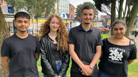 Zain, who has short dark hair and is wearing a black t-shirt, Amelia, who has long brown curly hair and is wearing a leather jacket, Samir, who has short hair and a moustache and goatee, and is wearing a black polo, and Safi, who has tied back black hair and is wearing a black t-shirt, stand together smiling outside. 