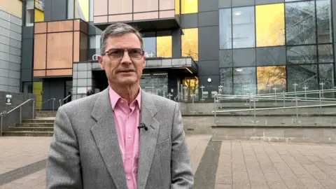 A man wearing glasses, with greying hair, a grey jacket and a pink shirt stands in front of County Hall in Trowbridge