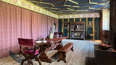 A roof with pink curtains along the left wall. The ceiling is blue and hold. There is a long dark wood table in the centre of the room. The carpet is light brown.