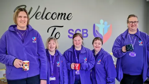 Supported Community Business A group of people wearing purple fleece tops are standing against a grey wall which has writing on it which reads "welcome to SCB". Three members of the group are holding mugs, all are smiling for photograph.