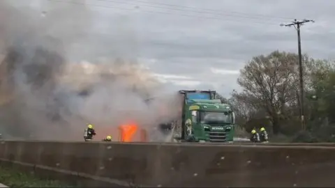 Alana Webb Four firefighters, in protective clothing and yellow helmets, trying to extinguish a fire on the M5. At the bottom of the picture is the motorway barrier, and above is a green lorry with flames coming out of the side. 