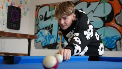 A teenager plays pool on a blue table. The camera is positioned in front of a ball they are about to attempt to pot. The teenager, who is wearing a black and white hoodie has brown hair and is smiling at the camera. A wall behind is covered in graffiti.