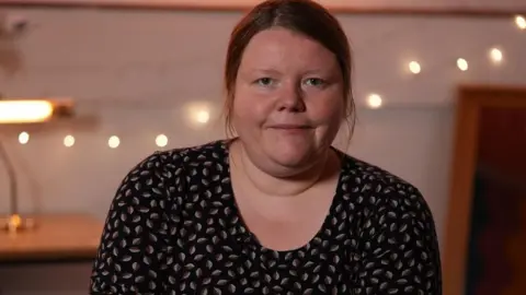 Helen is wearing a black, patterned dress. She has long brown, tied-back hair.